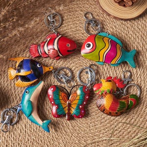May include: A selection of vibrant, handmade keyrings in diverse designs. The keyrings showcase a red and white clownfish, a rainbow-hued fish, a blue and yellow fish, a turquoise dolphin, a butterfly, and a rooster.
