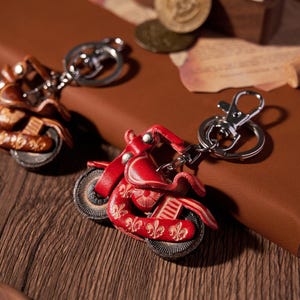 May include: Two motorcycle keychains. One is red with silver accents, the other is brown. Both have intricate details and silver key rings. The keychains are on a brown leather surface with coins and a small wooden box in the background.