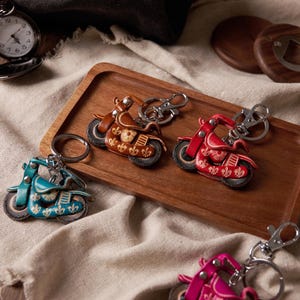 Op de afbeelding: Leren motorfiets sleutelhangers in teal, bruin en rood met zilveren sleutelringen. De sleutelhangers liggen op een houten dienblad met een zakhorloge en flesopener op de achtergrond. De motorfiets sleutelhangers hebben decoratieve details.