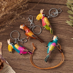 Könnte beinhalten: Vier papageienförmige Schlüsselanhänger aus Leder in leuchtenden Farben wie Rot, Gelb, Grün, Blau und Pink. Jeder Vogel zeigt ein detailliertes Flügelmuster, einen markanten Schnabel und ein kreisförmiges Auge. Drei haben Metallringe und Clips.