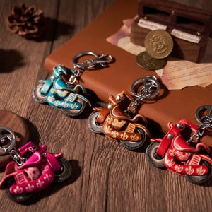 Op de afbeelding: Een verzameling motorfiets sleutelhangers in verschillende kleuren, waaronder teal, bruin, roze en rood. Elke sleutelhanger heeft ingewikkelde reliëfdetails en een zilverkleurige sleutelring. De sleutelhangers worden gepresenteerd op een houten oppervlak.