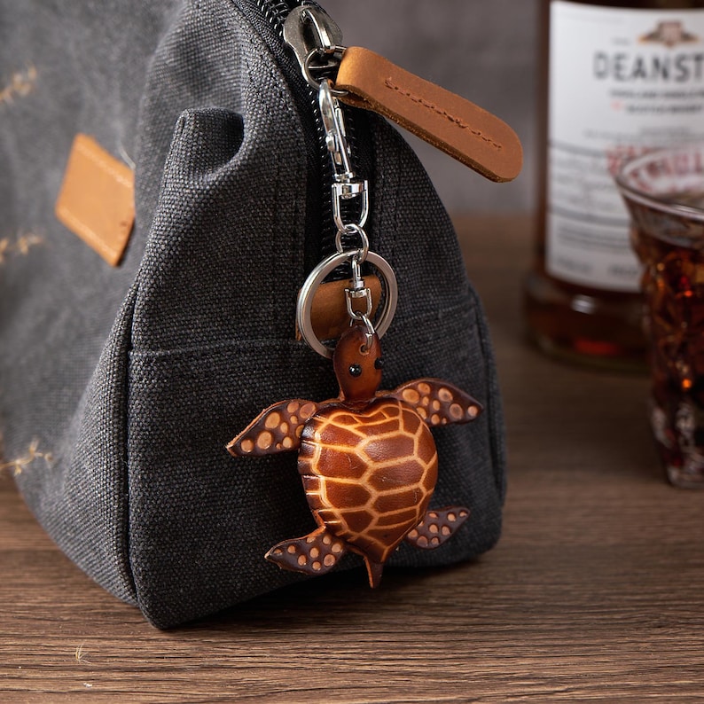May include: A brown leather turtle keychain hanging from a silver zipper pull on a gray canvas bag. The turtle has a detailed, textured design. A brown leather pull tab is attached to the zipper. A bottle of liquor is in the background.