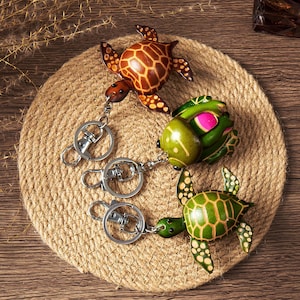 May include: Three handcrafted turtle keychains in shades of brown, green, and yellow, each with a unique shell pattern. The keychains are attached to silver metal clasps and are displayed on a round, woven mat.