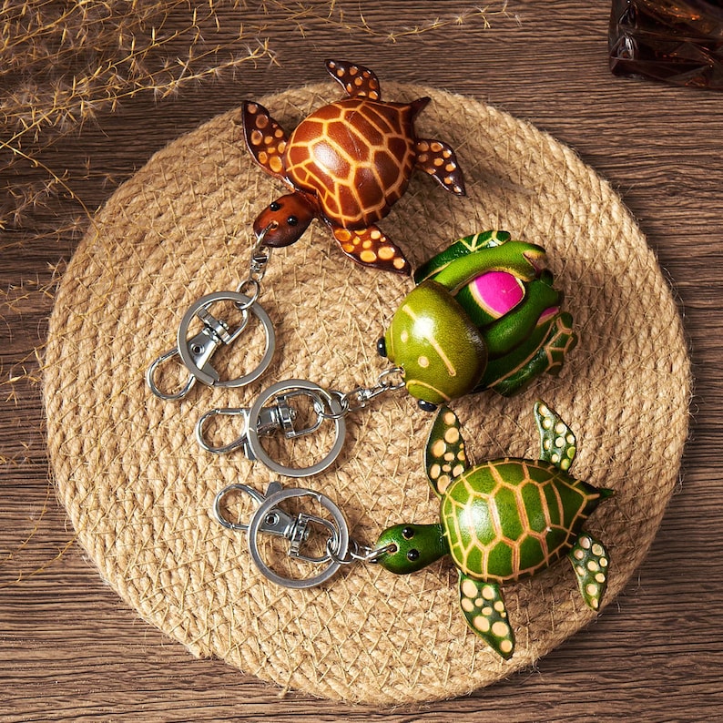 May include: Three handcrafted turtle keychains in shades of brown, green, and yellow, each with a unique shell pattern. The keychains are attached to silver metal clasps and are displayed on a round, woven mat.