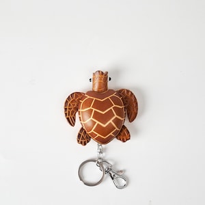 May include: A brown leather turtle keychain with a silver keyring and clasp. The turtle has a detailed shell pattern and a textured appearance. The keychain is designed to resemble a sea turtle.
