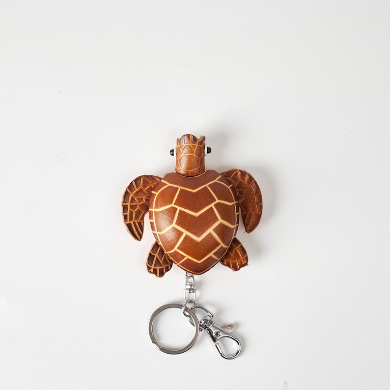 May include: A brown leather turtle keychain with a silver keyring and clasp. The turtle has a detailed shell pattern and a textured appearance. The keychain is designed to resemble a sea turtle.