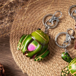 May include: A green leather frog keychain with a silver keyring. The frog has a pink belly and yellow accents. The keychain is on a brown, textured surface.