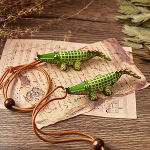 Pode incluir: Dois pingentes em forma de crocodilo verde com um padrão de grade e detalhes em branco, cada um preso a um fino cordão de couro bege. Os crocodilos apresentam pequenos olhos brancos e estão conectados pelos cordões com contas de madeira.