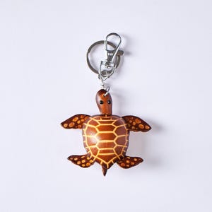 May include: A brown leather turtle keychain with a silver-colored clasp. The turtle has a detailed shell pattern and small, round eyes. The keychain is a decorative accessory.