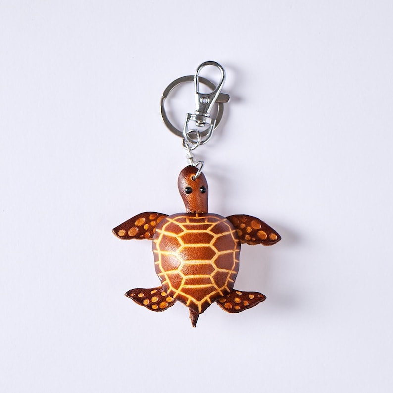 May include: A brown leather turtle keychain with a silver-colored clasp. The turtle has a detailed shell pattern and small, round eyes. The keychain is a decorative accessory.