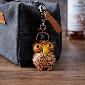 Może przedstawiać: Skórzany brelok do kluczy w kształcie sowy z brązowymi, żółtymi i zielonymi detalami wisi przed szarą płócienną torbą. Sowa ma duże żółte oczy i żółty dziób. Brelok ma srebrny zatrzask i brązowy skórzany pasek.