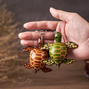 May include: Two leather turtle keychains. One is brown with a geometric pattern, the other is green with a striped pattern. Both have small, dark eyes and are attached to a silver keychain ring.