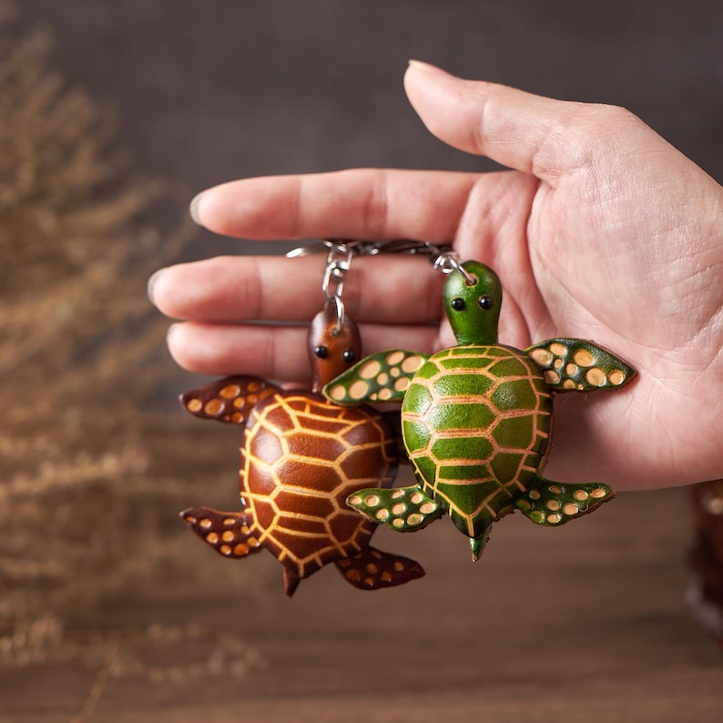 May include: Two leather turtle keychains. One is brown with a geometric pattern, the other is green with a striped pattern. Both have small, dark eyes and are attached to a silver keychain ring.