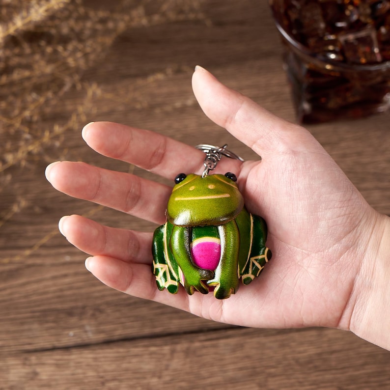 May include: A green and pink frog keychain with a silver keyring. The frog has black eyes and a pink belly. The keychain is made of leather and has a detailed design.