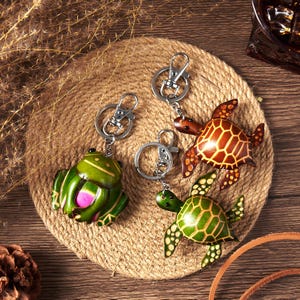 May include: Leather keychains featuring a green frog and two turtles. The frog has a pink belly, and the turtles have brown and green shells. Each keychain has a silver-colored clasp and ring, set on a woven, circular mat.