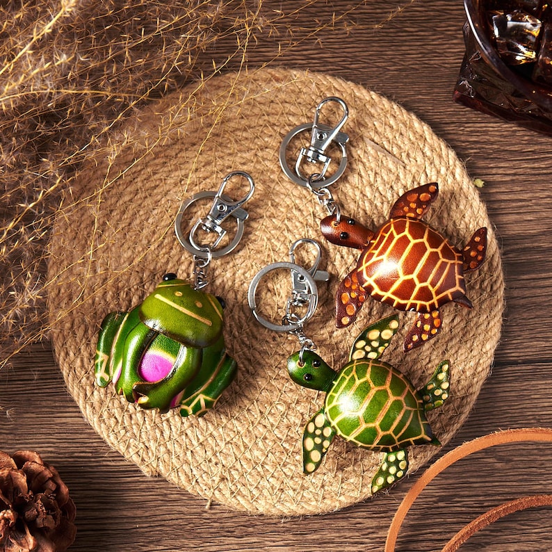 May include: Leather keychains featuring a green frog and two turtles. The frog has a pink belly, and the turtles have brown and green shells. Each keychain has a silver-colored clasp and ring, set on a woven, circular mat.
