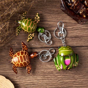 May include: Four leather keychains: two turtles and two frogs. The turtles are brown and green, with detailed patterns. The frogs are green with pink accents. Each keychain has a silver-colored clasp.