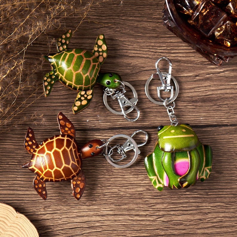 May include: Four leather keychains: two turtles and two frogs. The turtles are brown and green, with detailed patterns. The frogs are green with pink accents. Each keychain has a silver-colored clasp.