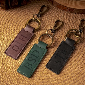 Peut inclure: Trois porte-clés personnalisés en cuir marron, vert et noir. Chaque porte-clés comporte une étiquette rectangulaire en cuir avec des initiales gravées et un fermoir et un anneau en métal. Les porte-clés sont présentés sur une surface tissée texturée.