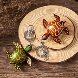May include: Two leather turtle keychains. One is green with a light green pattern, the other is brown with a darker brown pattern. Both have silver key rings and are on a wooden surface.