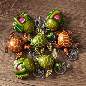 May include: A collection of handcrafted turtle keychains. The keychains feature detailed turtle designs in shades of green, brown, and pink, with silver key rings. The turtles have textured shells and flippers, and are arranged on a wooden surface.