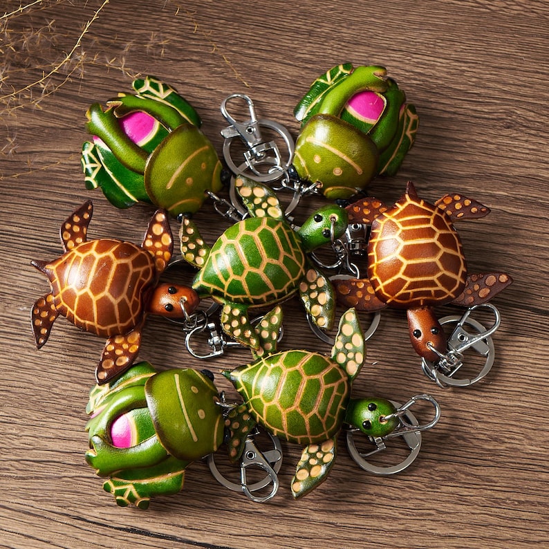 May include: A collection of handcrafted turtle keychains. The keychains feature detailed turtle designs in shades of green, brown, and pink, with silver key rings. The turtles have textured shells and flippers, and are arranged on a wooden surface.