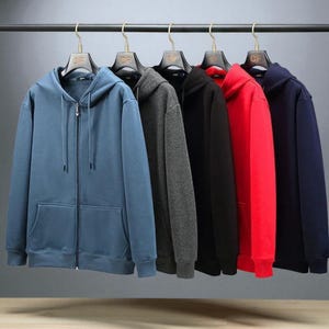 Può includere: Cinque felpe con cappuccio e zip di vari colori sono appese a un'asta di metallo. I colori includono blu, grigio, nero, rosso e blu navy. Ogni felpa ha un cappuccio e una tasca frontale. Le cerniere sono argentate.
