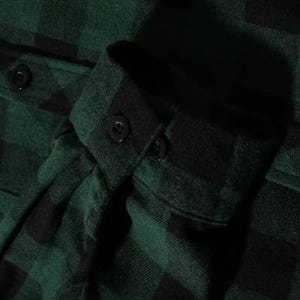 May include: A long-sleeved shirt featuring a dark green and black buffalo plaid pattern. The garment includes a structured cuff with a single black button closure and a matching black button on the sleeve placket, crafted from a soft fabric.