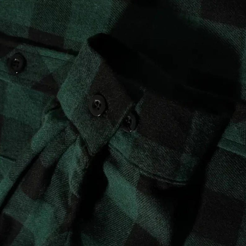 May include: A long-sleeved shirt featuring a dark green and black buffalo plaid pattern. The garment includes a structured cuff with a single black button closure and a matching black button on the sleeve placket, crafted from a soft fabric.