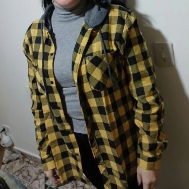 May include: A long-sleeved button-down shirt featuring a yellow and black buffalo plaid pattern, a gray attached hood with matching drawstrings, and a single chest pocket. The shirt is worn over a gray mock-neck long-sleeved base layer garment.