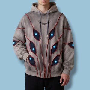 Könnte beinhalten: Grauer Kapuzenpullover mit einem einzigartigen Design mit roten Nähten und dunkelblauen, augenförmigen Akzenten mit leuchtend blauen Zentren. Der Hoodie hat eine Kapuze mit Kordelzug und eine kleine Grafik in der Nähe der Kapuze.
