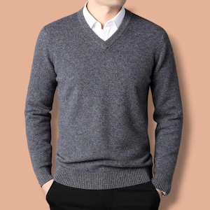 May include: A grey V-neck sweater with long sleeves, worn over a white collared shirt. The person is wearing black trousers. The background is a solid peach colour. The sweater has a ribbed hem and cuffs.