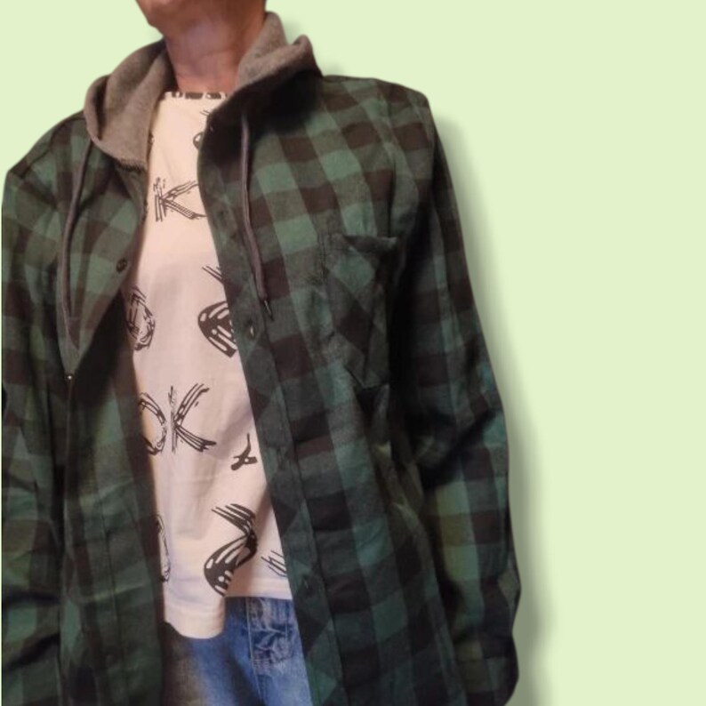 May include: A long-sleeved button-up shirt featuring a dark green and black buffalo plaid pattern, paired with a grey hooded collar and drawstrings. Underneath, a white t-shirt displays a black graphic print of abstract shapes and lines.