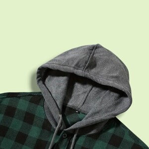 May include: A long-sleeve shirt featuring a dark green and black buffalo plaid pattern. The garment includes an attached heather gray hood made of a soft fabric, complete with matching gray drawstrings and a small black size tag labeled M.