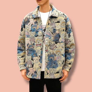 Op de afbeelding: Een jas met knopen met een herhaald patroon van teddyberen in verschillende kleuren, waaronder blauw, beige en bruin. De jas heeft een kraag en twee voorzakken. De persoon draagt eronder een wit t-shirt.