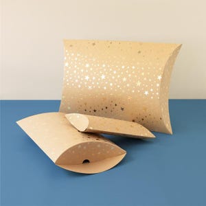 Könnte beinhalten: Drei beige Kissenboxen mit einem goldenen Sternmuster. Eine Box steht aufrecht, und zwei sind offen und liegen flach. Die Boxen befinden sich auf einer blauen Oberfläche.
