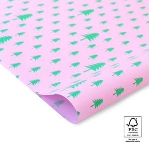 Könnte beinhalten: Ein Bogen hellrosa Geschenkpapier mit einem sich wiederholenden Muster aus kleinen, leuchtend grünen Tannenbäumen, die in verschiedene Richtungen ausgerichtet sind. Unten rechts befindet sich ein kleines weißes Quadrat mit dem FSC-Logo.