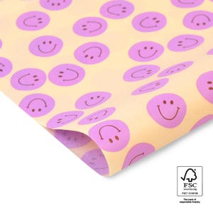 Könnte beinhalten: Geschenkpapier mit einem sich wiederholenden Muster aus lila Smiley-Gesichtern auf hellgelbem Hintergrund. Das Papier ist gefaltet, wodurch seine Textur sichtbar wird. Das FSC-Logo ist in der Ecke zu sehen.