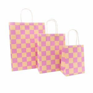 Könnte beinhalten: Drei Geschenkpapierbeutel in verschiedenen Größen, jeweils mit einem rosa- und hellorangenfarbenen Schachbrettmuster. Die Beutel haben weiße gedrehte Papiergriffe und stehen vor einem weißen Hintergrund.
