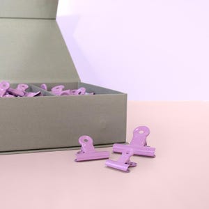 Könnte beinhalten: Eine Sammlung von lavendelfarbenen Büroklammern, einige in einer grauen Box und andere auf einer rosa Oberfläche verstreut. Die Klammern haben ein klassisches Design mit Federmechanismus. Die Box ist geöffnet und zeigt weitere Klammern.