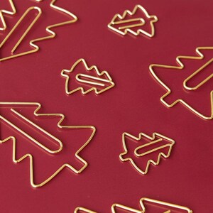 Könnte beinhalten: Eine Sammlung goldfarbener Metall-Büroklammern in Form stilisierter Weihnachtsbäume. Die Drahtklammern sind in einem verstreuten Muster vor einem einfarbigen, dunkelroten Hintergrund angeordnet und zeigen ihre markanten dreieckigen Baum-Silhouetten.