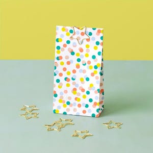 Könnte beinhalten: Eine weiße Geschenktüte mit buntem Tupfenmuster, gekrönt von einer goldfarbenen sternförmigen Klammer. Mehrere goldfarbene, sternförmige Büroklammern liegen um die Tüte herum, was ein festliches Thema ergibt.