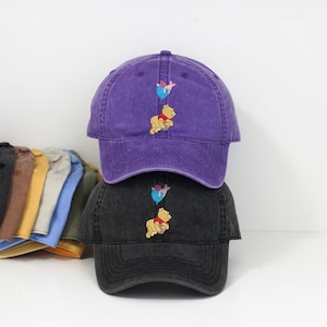 May include: Two baseball caps, one purple and one black, stacked on top of each other. Each cap features an embroidered design of a yellow bear with a blue balloon. Other caps in various colors are visible in the background.