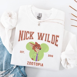 May include: White crewneck sweatshirt featuring the text "NICK WILDE" in brown lettering. Below is an embroidered image of Nick Wilde from Zootopia, with the text "EST. 2016 ZOOTOPIA". A silver necklace with a pendant is also visible.