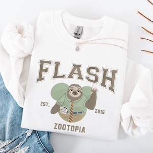 May include: White sweatshirt featuring an embroidered design of Flash from Zootopia. The design includes the word "FLASH" in a collegiate font, a cartoon sloth character, and the text "ZOOTOPIA EST. 2016". A silver necklace with a pendant is draped on the sweatshirt.