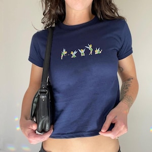 May include: Navy blue crop top with short sleeves. The shirt features an embroidered design of several fairies in various poses across the chest. The fairies are depicted in green, yellow, and white. The shirt is a fitted style.