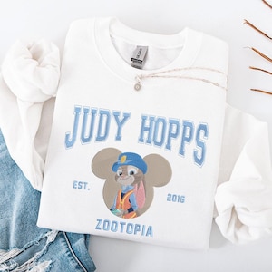 May include: White sweatshirt featuring the text "JUDY HOPPS" in light blue, with an image of Judy Hopps from Zootopia. The design includes the text "EST. 2016" and "ZOOTOPIA". A silver necklace with a small pendant is also visible.