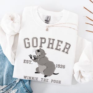 May include: White sweatshirt with the word "GOPHER" in gray block letters above a cartoon gopher illustration. The text "EST. 1926" and "WINNIE THE POOH" are also printed on the sweatshirt. A silver necklace with a small pendant is on the sweatshirt.
