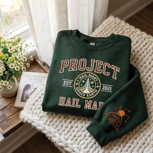 May include: A dark green sweatshirt with the words "PROJECT HAIL MARY" in white and brown lettering. The sweatshirt features a circular graphic with the words "HAIL MARY GRACE EARTH" and the year "2021". The sleeve has a graphic of an insect and the words "YOU SLEEP I WATCH".