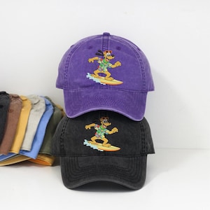 May include: Two baseball caps, one purple and one black, are stacked. Each cap features an embroidered cartoon dog surfing on a yellow surfboard. The caps are made of a soft, textured fabric and have a curved brim.